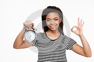 Afro american woman holding alarm clock and showing ok gesture