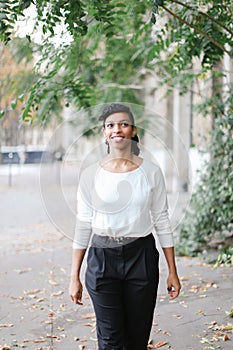 Afro american woman with ponytail walking in town near green trees and building, wearing white blouse.