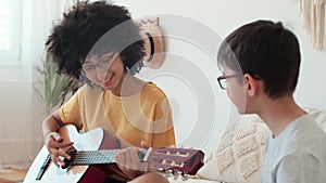 Afro american woman teaching boy to play guitar while sitting on bed at home