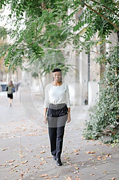 Afro american woman walking in town near green trees and building, wearing white blouse.