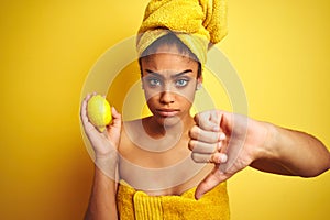 Afro american woman wearing towel after shower holding lemon over isolated yellow background with angry face, negative sign