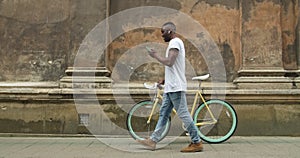 Afro American Young Man in Earphones Using his Modern Mobile Phone and Pushing Stylish Thin Tire Bike while Going along