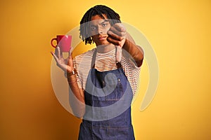 Afro barista man with dreadlocks drinking cup of coffee over isolated yellow background with angry face, negative sign showing