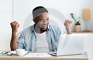 Afro businessman celebrating success with raised arms looking to laptop