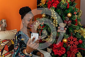 Afro-descendant Colombian woman sitting on the sofa next to the Christmas tree with her cell phone