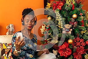 Afro-descendant Colombian woman sitting on the sofa next to the Christmas tree listening to music with headphones on her cellphone