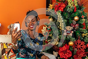 Afro-descendant Colombian woman sitting on the sofa next to the Christmas tree listening to music with headphones on her cellphone