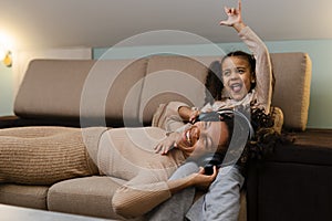 Afro girl and her little daughter in headphones is listening to music and smiling while lying on couch at home