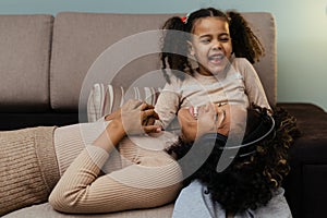 Afro girl and her little daughter in headphones is listening to music and smiling while lying on couch at home