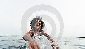 Afro kid splashing water on camera while swimming inside the ocean during summer vacation - African child having fun playing on