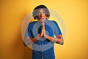 Afro man with dreadlocks wearing casual denim shirt standing over isolated yellow background begging and praying with hands