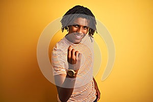 Afro man with dreadlocks wearing striped t-shirt standing over isolated yellow background Beckoning come here gesture with hand