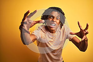 Afro man with dreadlocks wearing striped t-shirt standing over isolated yellow background Shouting frustrated with rage, hands