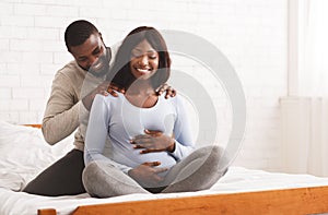 Afro man massaging his pregnant wife, sitting on bed