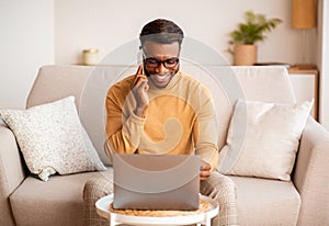 Afro Man Using Laptop Having Pleasant Phone Conversation At Home