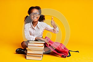 Afro Schoolgirl Gesturing Thumbs-Up Sitting At Book Stack, Studio Shot