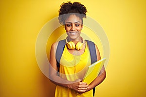 Afro woman using backpack and headphones holding notebook over isolated yellow background with a happy face standing and smiling