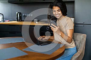 Afro beautiful woman using laptop and mobile phone while having breakfast in modern kitchen