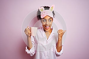 Afro woman wearing pajama and mask drinking a cup of coffee over isolated pink background pointing and showing with thumb up to