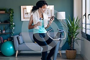 Afro young fitness girl using mobile phone while training on exercise bike at home