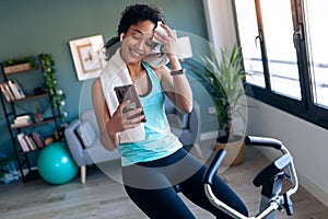 Afro young fitness girl using mobile phone while training on exercise bike at home