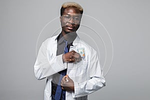 Afroamerican doctor putting dollars money in his pocket isolated on gray background