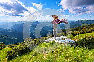 Against the background of beautiful mountain scenery with sun rays and cloudy sky the girl is staying in long dress, straw hat.