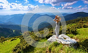 Against the background of beautiful mountain scenery with sun rays and cloudy sky the girl is staying in long dress, straw hat.