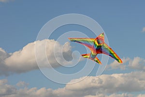 Against a background of blue sky with white clouds flies a bright kite in the form of a rainbow airplane. Summer games.