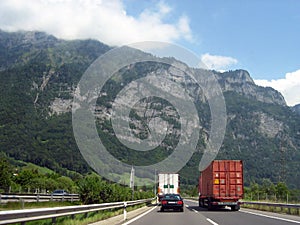 Against the background of a huge forest-covered mountain, trucks and cars drive along the road in perspective