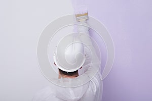 Against the background of a painted wall, the hand of a worker in a white protective helmet, holding a brush, rear view