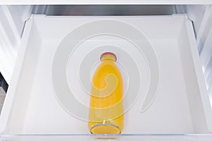 Against the background of a white refrigerator drawer, one glass bottle with orange juice, close-up