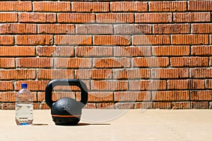 Against a brick wall, close-up of a black sports kettlebell and a bottle of water, there is a place for inscription