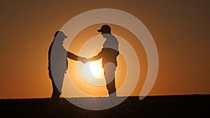 Against the setting sun over the field, the silhouettes of two farmers shake hands.