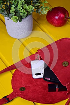 Against a yellow wooden background, a woman's leather red handbag is open, a wallet, a mobile phone and lipstick peep