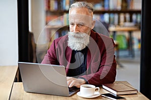 Aged businessman using laptop at cafe, drinking coffee