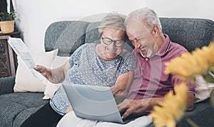 Aged couple at home paying bills on line with laptop and laughing a lot having fun together. Happiness and elderly lifestyle. Man