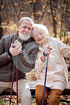 Aged couple resting while sitting on bench with nordic walking poles, smiling