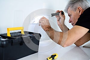 Aged electrician, repairman in uniform working, examining socket in the kitchen using flashlight. Repair service concept