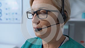 Aged female medical worker in headset typing on laptop, online consultation