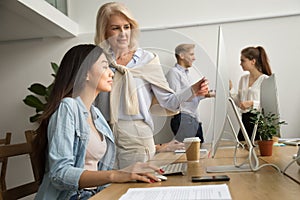 Aged female mentor helping asian girl intern explaining computer