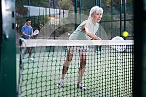 Aged female paddle tennis player performing backhand on outdoor court