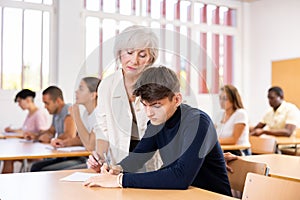 Aged female teacher helping guy student solving problem at lesson