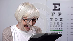 Aged lady in eyeglasses smiling and reading from tablet, focusing vision, health