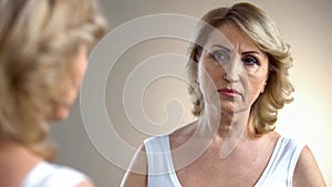 Aged lady looking in mirror on her wrinkled face, thinking about plastic surgery