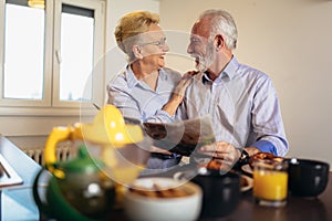 Aged loving couple reading newspaper at home