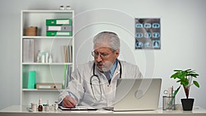 Aged male doctor reviews the patient's analyzes on a laptop and makes notes in his medical history. Doc in white medical