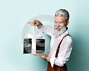 Aged male in white shirt, brown pants and suspenders, bracelet. He holding silver gift box, posing sideways on blue background