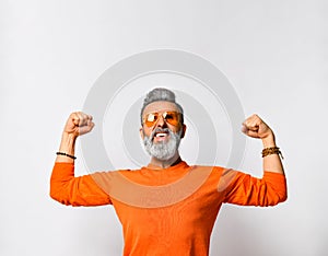 Aged man in orange sweater and sunglasses, bracelets. Clenched fists, showing his muscles, posing isolated on white. Close up