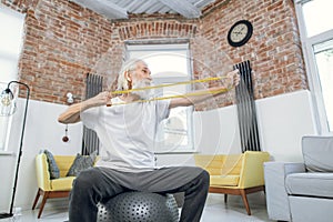 Aged man sitting on ball and training arms with rubber band